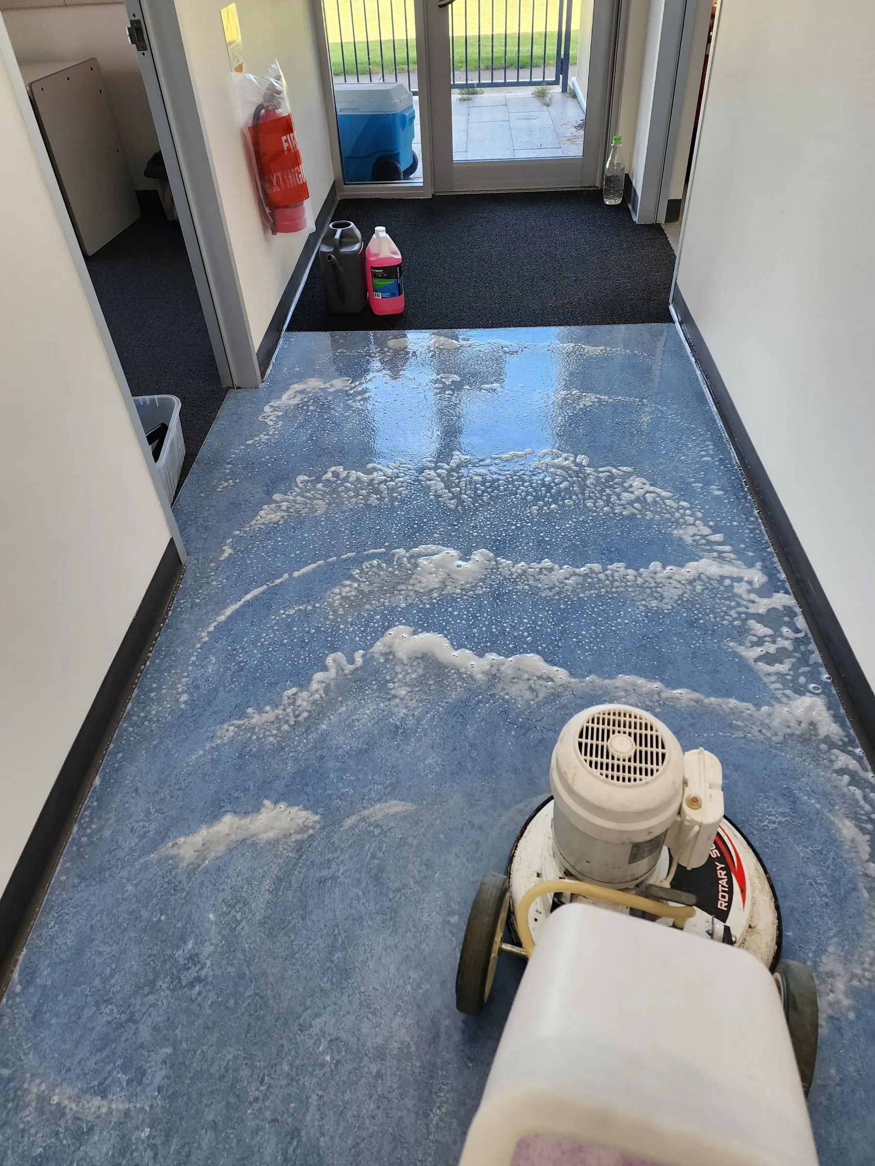 Rotary floor machine scrubbing a blue vinyl corridor floor, foam on the surface