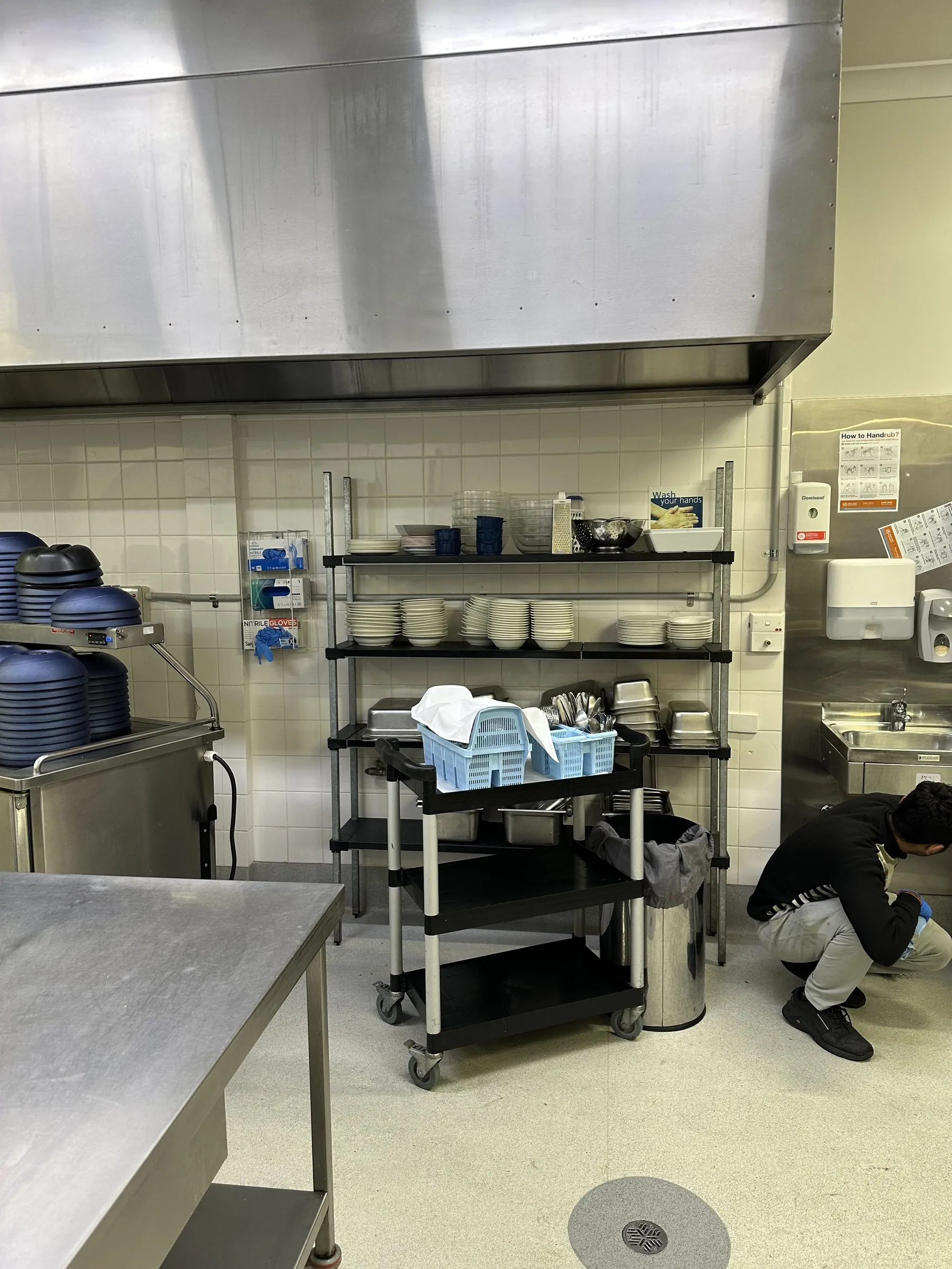 Person cleaning low under a stainless bench beside a trolley