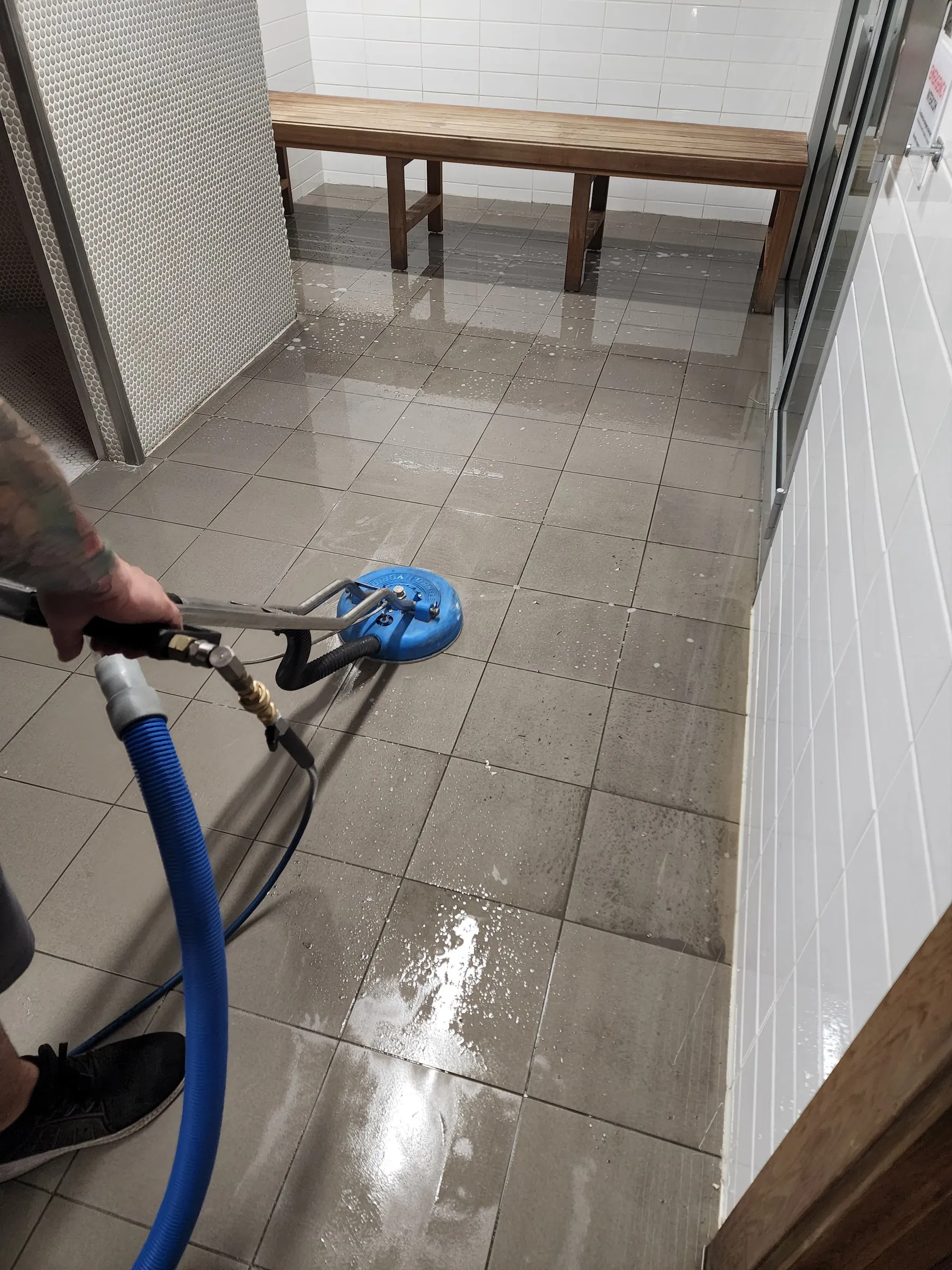 Spinner wand scrubbing grey floor tiles in a corridor beside a bench